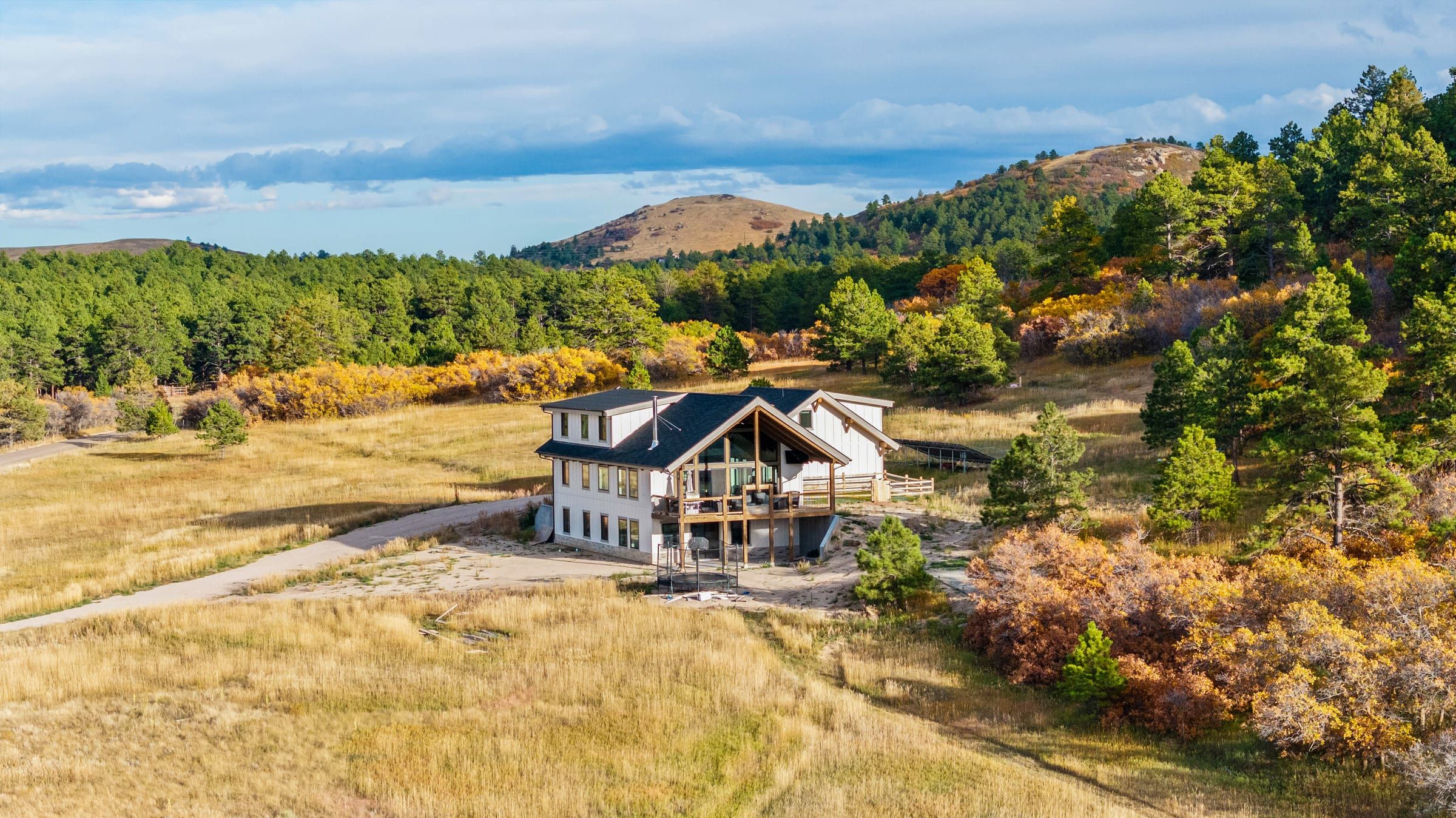 Aerial view of a custom home with a timber-framed gable deck, set among pines and autumn scrub oak below a hill