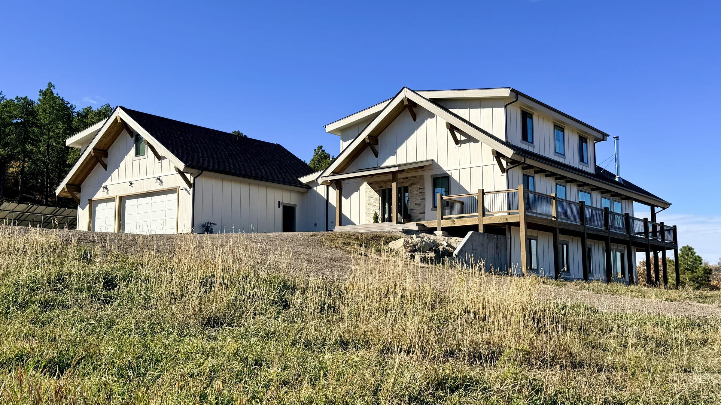 White board-and-batten custom home with timber-bracketed gables, a covered deck and an attached two-car garage on a grassy hillside under a clear blue sky